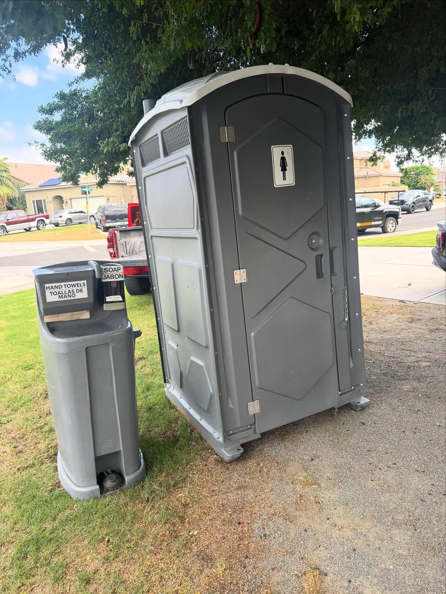 Gray deluxe portable toilet beside a freestanding handwashing sink on a lawn